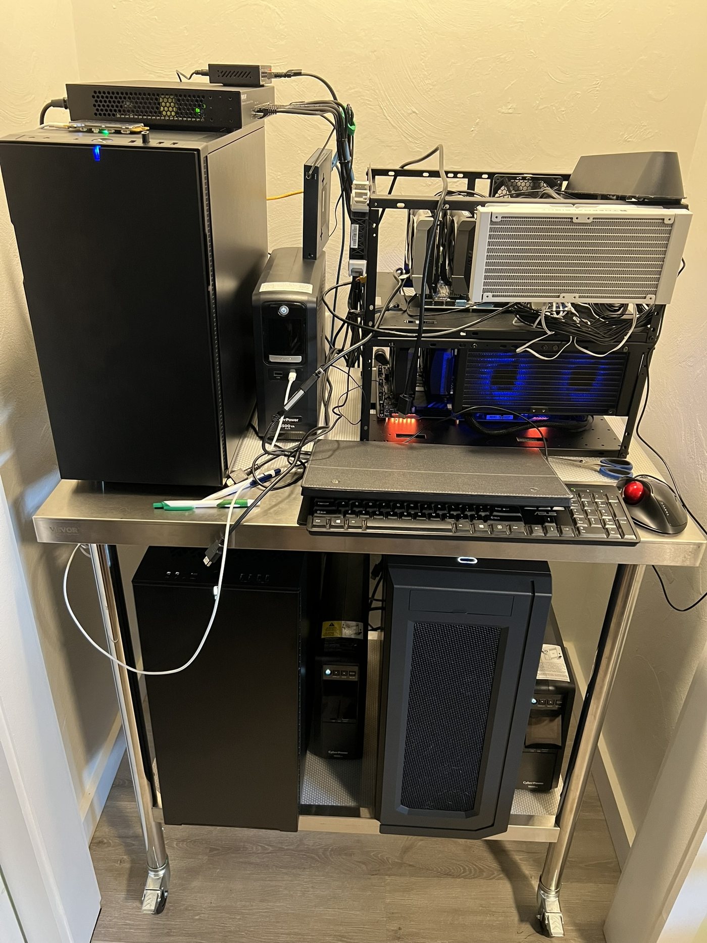 Home hive research cluster: black workstations stacked on a stainless steel cart with UPS battery backup, networking gear, and an open-frame machine with blue cooling fans.